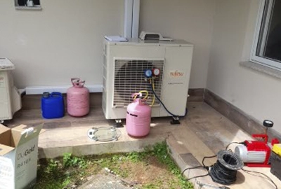 Equipamentos e cilindros de gás refrigerante organizados ao lado de unidade condensadora de ar-condicionado Fujitsu durante procedimento de carga de gás em área externa.