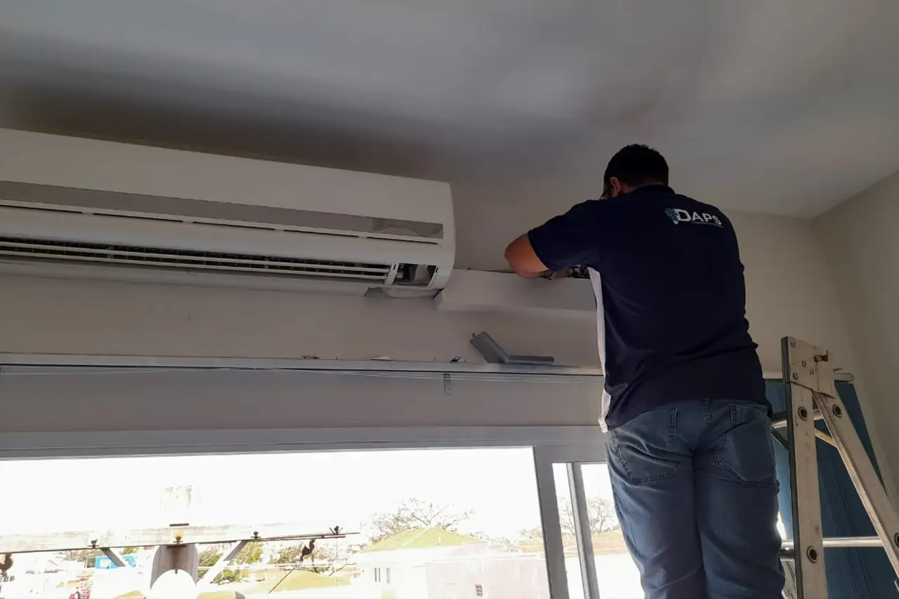 Técnico da DAPS Climatização realizando manutenção de ar-condicionado split residencial próximo ao teto, em ambiente com janela de vidro.