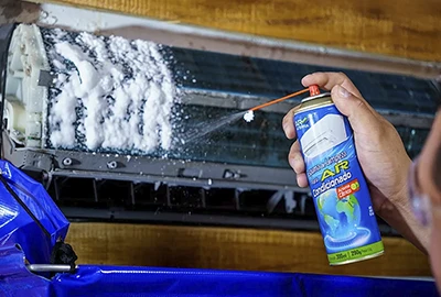 Mão aplicando espuma de higienização em spray diretamente na serpentina da unidade interna de ar-condicionado split, com capa protetora instalada.