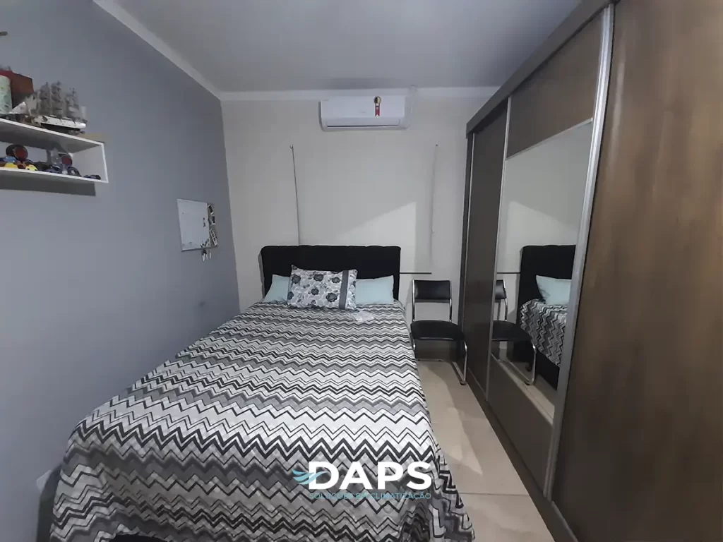 Quarto com ar-condicionado instalado na parede, cama com colcha padrão geométrico, espelho grande, prateleiras e iluminação natural, demonstrando a climatização de ambiente residencial.