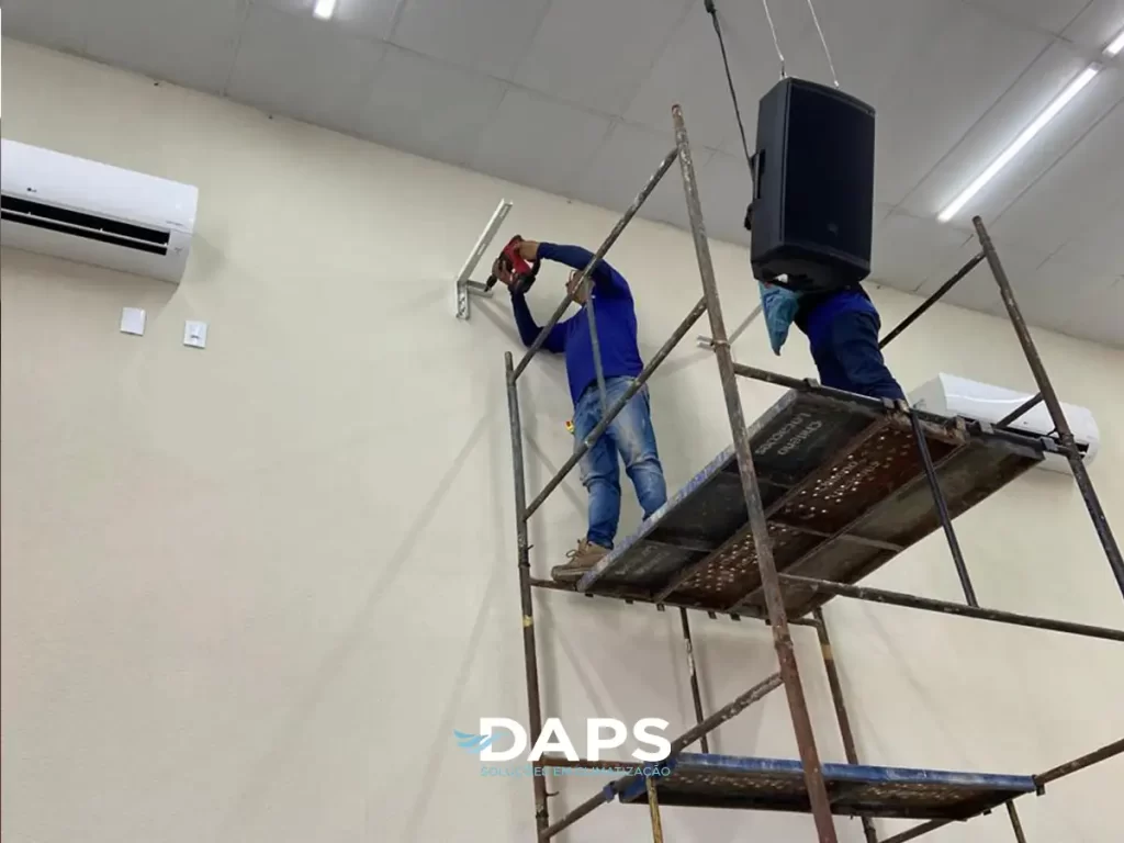 Dois técnicos em uniforme da DAPS trabalham em andaime metálico para instalar unidade de ar-condicionado em parede interna de ambiente em Campinas, demonstrando trabalho profissional em altura.