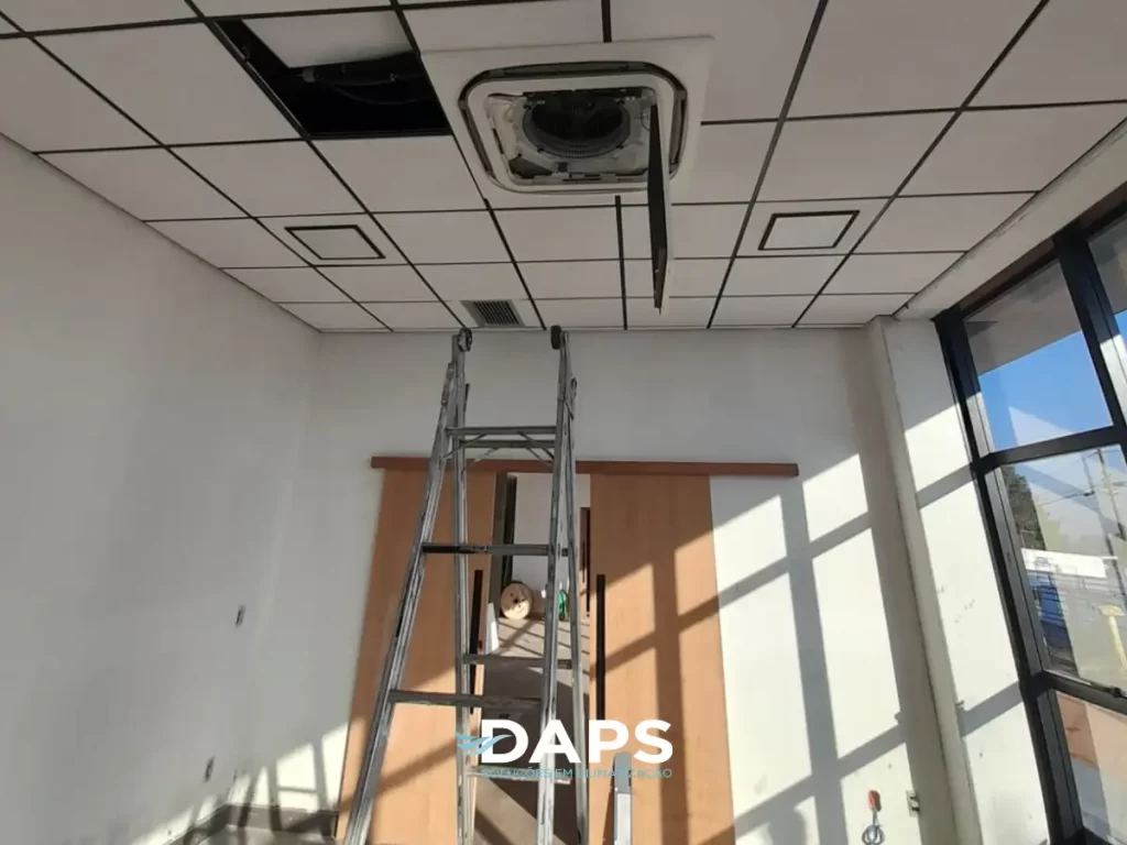 Técnico realizando instalação de ar-condicionado tipo cassete embutido em forro de gesso em ambiente comercial, utilizando escada para acesso ao teto, demonstrando trabalho profissional de climatização em prédio.