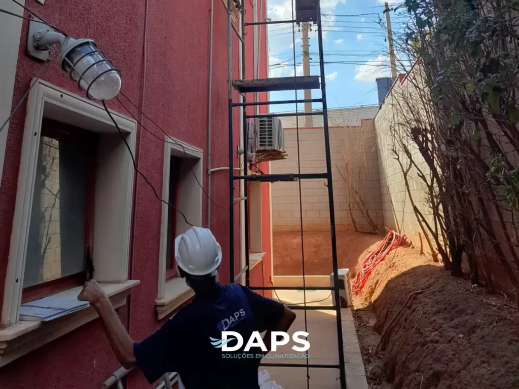 Técnico da DAPS realizando instalação de ar-condicionado em parede externa de edifício, com unidade externa sendo posicionada em estrutura metálica, demonstrando processo profissional de instalação.