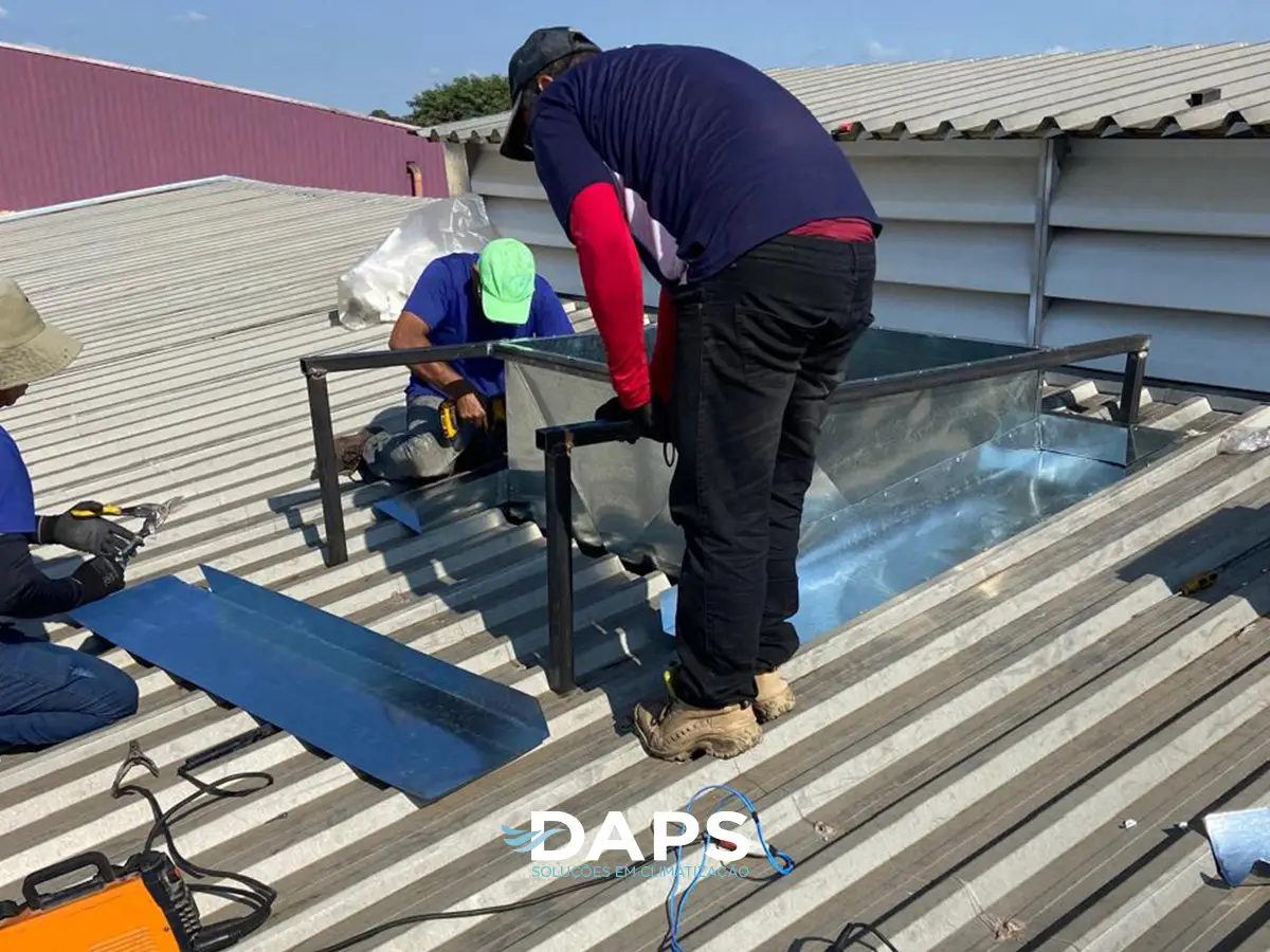 Dois técnicos DAPS em uniforme instalando ar-condicionado em cobertura de supermercado em Campinas, trabalho em altura com estrutura metálica, equipamento de climatização para grande área.