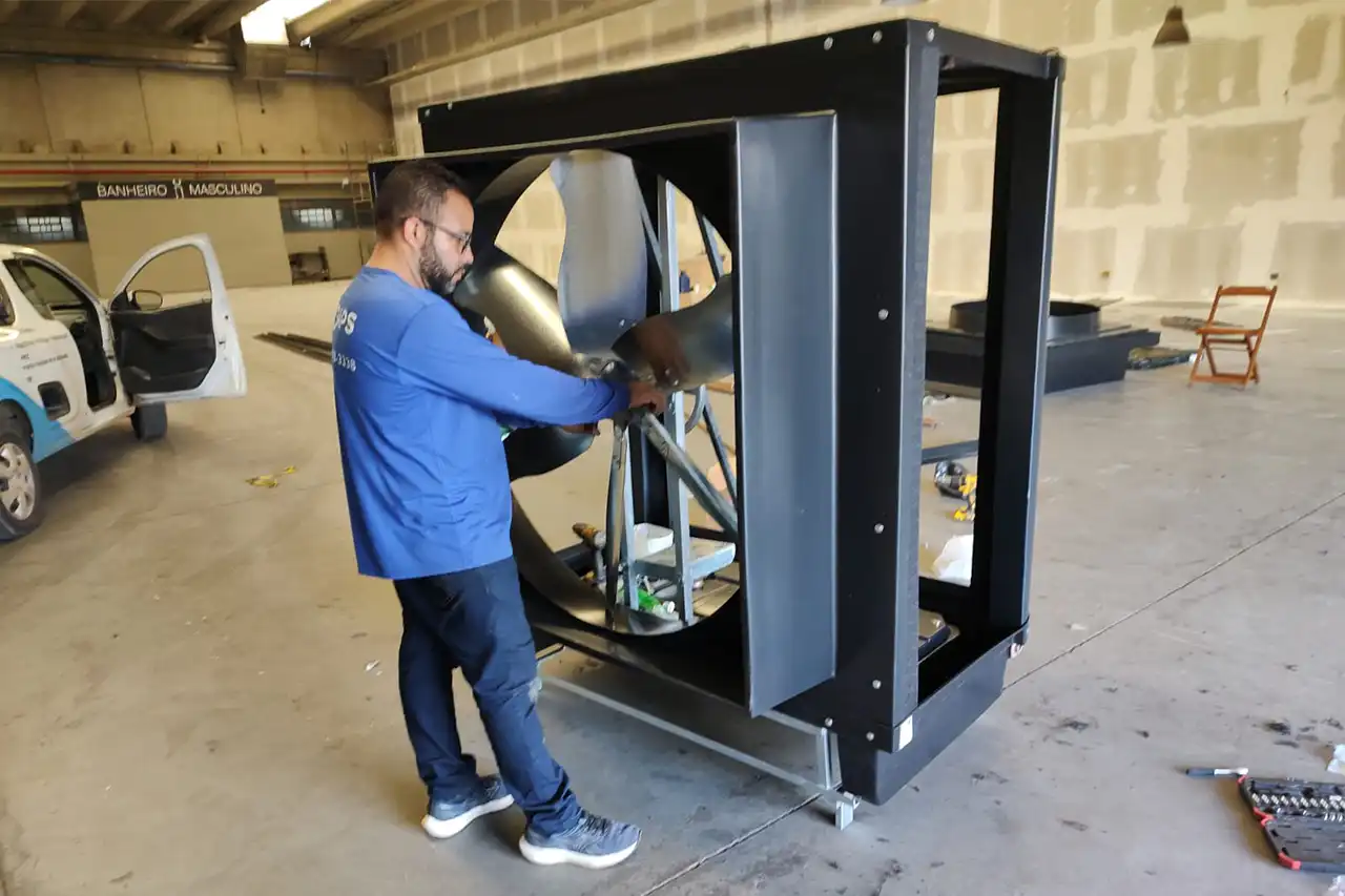Fotografia fotorrealista do interior de um amplo galpão industrial em Campinas, São Paulo, exibindo um sistema de climatização industrial evaporativo instalado. Placas de parede com 'DAPS Climatização | Instalação e Engenharia em Climatizador Industrial', 'Engenharia Certificada CREA-SP' e 'PMOC' são visíveis. Um técnico da DAPS uniformizado aponta para os dutos para um gestor. A linha do horizonte de Campinas é vista através das janelas. Foco no conforto térmico e engenharia certificada.